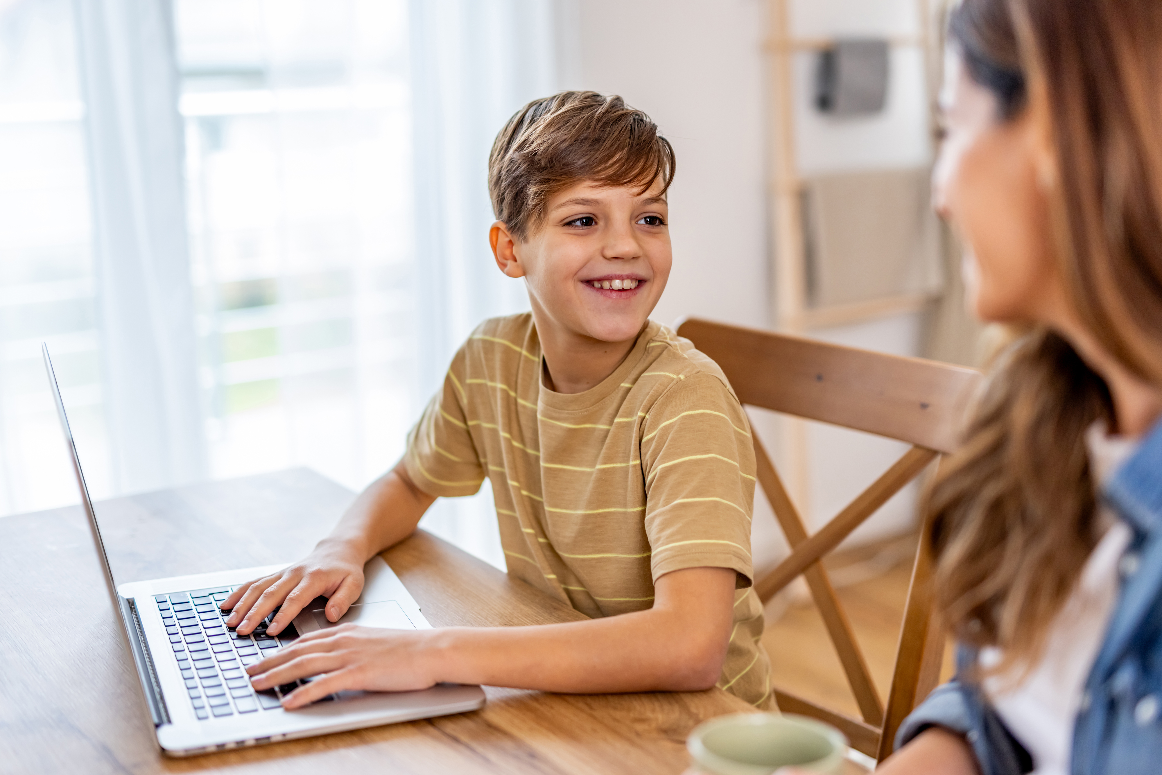 A boy smiling while using a laptop at a table with a parent nearby